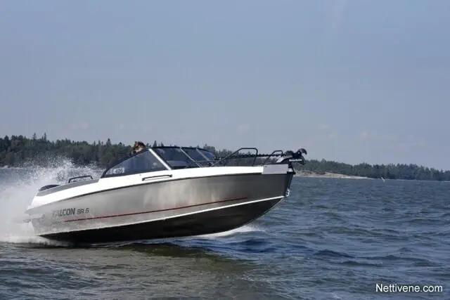 A sleek Falcon 98.6 boat speeds across the water, creating waves against a backdrop of trees and a clear sky.